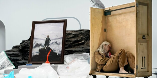 Eine Frau sitzt in einer Holzbox, daneben Caspar David Friedrichs Gemälde "Mann im Nebelmeer"