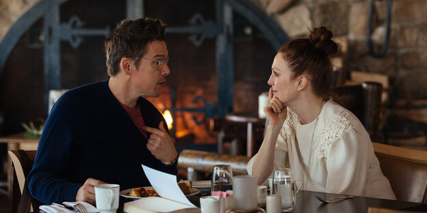 Ein Ethan Hawke und eine Julianne Moore sitzen sich in an einem Tisch beim Essen gegenüber, hinter ihnen lodert ein Feuer im Kamin. Ein Ethan Hawke und eine Julianne Moore sitzen sich in an einem Tisch beim Essen gegenüber, hinter ihnen lodert ein Feuer im Kamin.