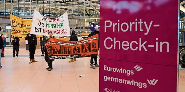 Ein violettes Priority-Check-In-Schild, daneben DemonstrantInnen Ein violettes Priority-Check-In-Schild, daneben DemonstrantInnen