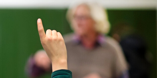 Die Hand einer Schülerin, die sich gerad meldet, im Hintergrund der Lehrer.