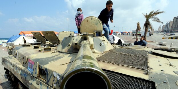 Zwei Kinder stehen auf einem Panzer. Im Hintergrund sind Häuserfassaden und eine Palme zu sehen.