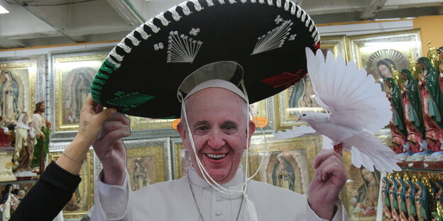 Eine Frau setzt dem Papst einen Mariachi-Hut auf. Er lacht in die Kamera und hält einen Spielzeugvogel in einer Hand Eine Frau setzt dem Papst einen Mariachi-Hut auf. Er lacht in die Kamera und hält einen Spielzeugvogel in einer Hand
