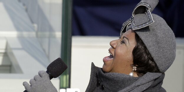 Aretha Franklin singt ins Mikro, trägt graue Mütze, Schal und Handschuhe Aretha Franklin singt ins Mikro, trägt graue Mütze, Schal und Handschuhe