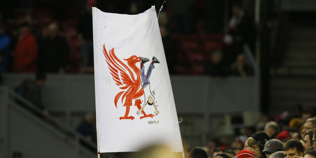 Ein Doppelhalter-Plakat in einem Fußballstadion: Darauf ist ein großer roter Vogel, der eine Person kopfüber im Schnabel hat. Die Person verliert Geld. Ein Doppelhalter-Plakat in einem Fußballstadion: Darauf ist ein großer roter Vogel, der eine Person kopfüber im Schnabel hat. Die Person verliert Geld.