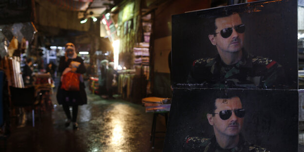Nächtliche Szene, rechts im Vordergrund zwei Bilder des syrischen Machthabers Assad mit Sonnenbrille, dahinter Markt Nächtliche Szene, rechts im Vordergrund zwei Bilder des syrischen Machthabers Assad mit Sonnenbrille, dahinter Markt