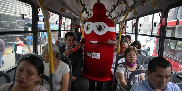 Ein Mensch in einem roten Kondomkostüm verteilt Kondome in einem Bus. Ein Mensch in einem roten Kondomkostüm verteilt Kondome in einem Bus.