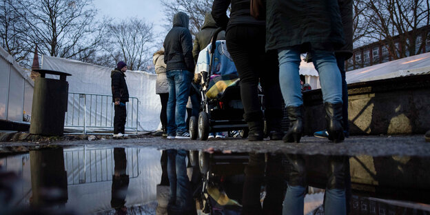 Menschen warten in einer Reihe vor einem Zelt. Neben ihnen ist eine Pfütze, in der sie sich spiegeln Menschen warten in einer Reihe vor einem Zelt. Neben ihnen ist eine Pfütze, in der sie sich spiegeln