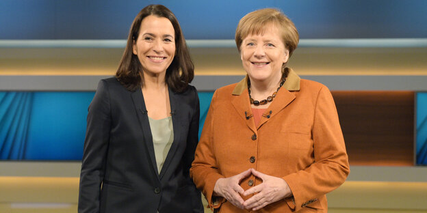 Anne Will steht lächelnd neben Angela Merkel, die mit ihren Händen eine Raute formt. Anne Will steht lächelnd neben Angela Merkel, die mit ihren Händen eine Raute formt.
