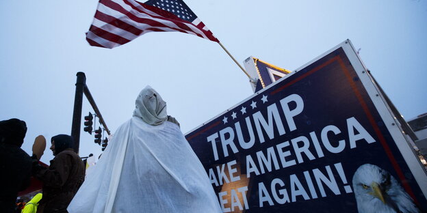 Ein Kritiker von Trump hat sich als Anhänger des Ku-Klux-Klans verkleidet und steht neben einem Schild „Trump make America great again!“. Ein Kritiker von Trump hat sich als Anhänger des Ku-Klux-Klans verkleidet und steht neben einem Schild „Trump make America great again!“.