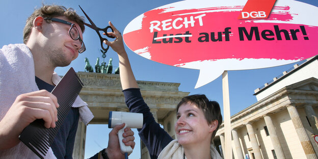Eine Aktivistin und ein Aktivist halten ein Transparent auf dem "Recht auf Mehr" steht, eine Schere und eine großen Kamm vor dem Brandenburger Tor Eine Aktivistin und ein Aktivist halten ein Transparent auf dem "Recht auf Mehr" steht, eine Schere und eine großen Kamm vor dem Brandenburger Tor