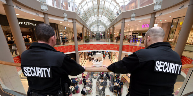 zwei Menschen in Schwarz mit Aufdruck "Security" auf dem Rücken blicken von einer Galerie runter in ein Einkaufszentrum zwei Menschen in Schwarz mit Aufdruck "Security" auf dem Rücken blicken von einer Galerie runter in ein Einkaufszentrum