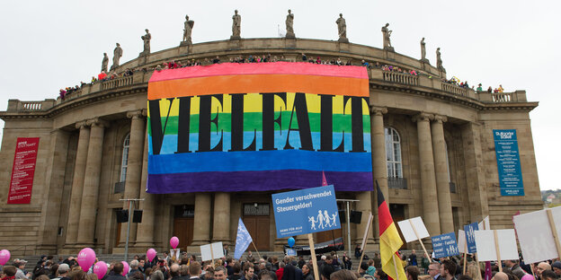 Gebäude mit Säulen, an dem ein riesiges Plakat in Regenbogenfarben mit der Aufschrift "Vielfalt" hängt, davor Menschen, einer davon mit einem Plakat auf dem steht: "Gender verschwendet Steuergelder" Gebäude mit Säulen, an dem ein riesiges Plakat in Regenbogenfarben mit der Aufschrift "Vielfalt" hängt, davor Menschen, einer davon mit einem Plakat auf dem steht: "Gender verschwendet Steuergelder"