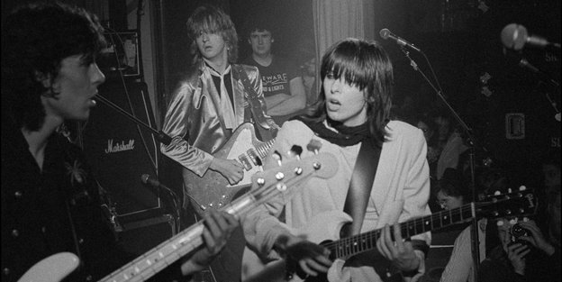 Das Schwarz-Weiß-Bild zeigt eine Frau mit Gitarre, die neben anderen Männern mit Gitarre steht.