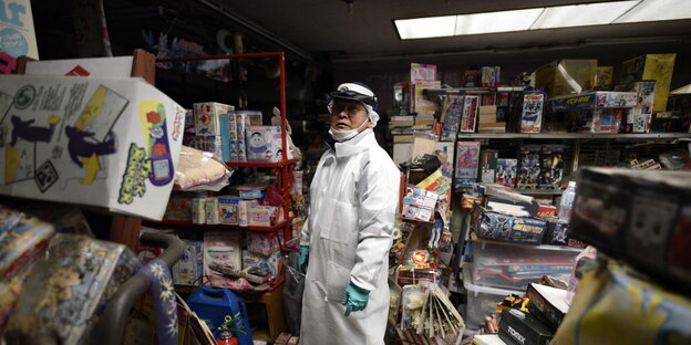 EIn MAnn mit Schutzkleidung steht in einem Laden zwischen Regalen mit unterschiedlichen Waren EIn MAnn mit Schutzkleidung steht in einem Laden zwischen Regalen mit unterschiedlichen Waren