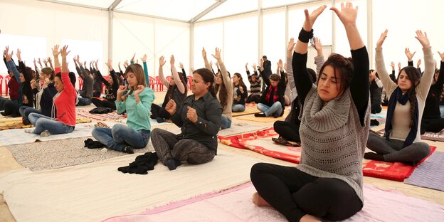 Frauen sitzen in einem Zelt im Yogasitz auf Matten und strecken die Arme nach oben. Frauen sitzen in einem Zelt im Yogasitz auf Matten und strecken die Arme nach oben.