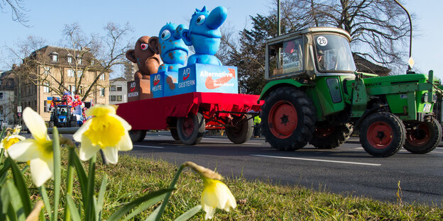 Ein Karnevalswagen stellt drei Figuren dar, die sich von blau nach braun verfärben. Er wird von einem grünen Traktor gezogen. Im Vordergrund stehen gelbe Blumen. Ein Karnevalswagen stellt drei Figuren dar, die sich von blau nach braun verfärben. Er wird von einem grünen Traktor gezogen. Im Vordergrund stehen gelbe Blumen.
