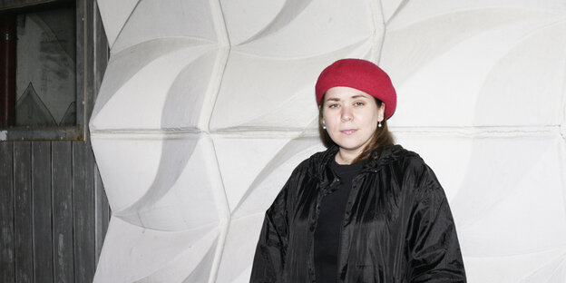 Stefanie Sargnagel in schwarzer Jacke und roter Mütze lehnt an einer grau-weißen Wand. Stefanie Sargnagel in schwarzer Jacke und roter Mütze lehnt an einer grau-weißen Wand.