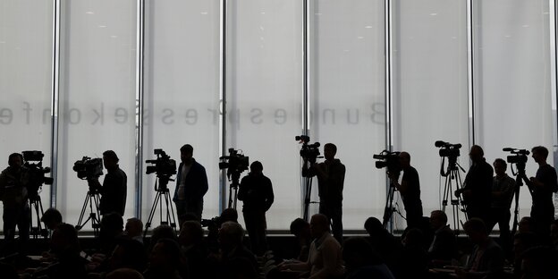 Journalisten mit Kameras stehen vor einer grau-weiße Wand.