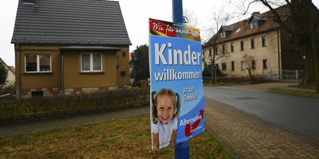 Ein AfD-Plakat, das Kinder willkommen heißt, hängt an einem blauen Pfahl, dahinter graubraune Häuser. Ein AfD-Plakat, das Kinder willkommen heißt, hängt an einem blauen Pfahl, dahinter graubraune Häuser.