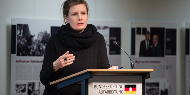 Eine Frau in dunkler Kleidung steht hinter einem Redepult, dahinter an der Wand hängen Ausstellungstafeln. Eine Frau in dunkler Kleidung steht hinter einem Redepult, dahinter an der Wand hängen Ausstellungstafeln.
