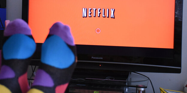 Zwei hochgelegte Füße mit bunten Socken vor Fernsehgerät, auf dem "Netflix" steht Zwei hochgelegte Füße mit bunten Socken vor Fernsehgerät, auf dem "Netflix" steht