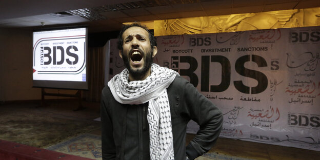 Ein Mann in dunkler Kleidung und Kufiya steht schreiend vor dem Schriftzug „BDS“ an einer Wand. Ein Mann in dunkler Kleidung und Kufiya steht schreiend vor dem Schriftzug „BDS“ an einer Wand.