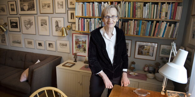 eine Frau steht am Tisch in einem Arbeits-/Wohnzimmer, an der Wand ein großes Bücherregal