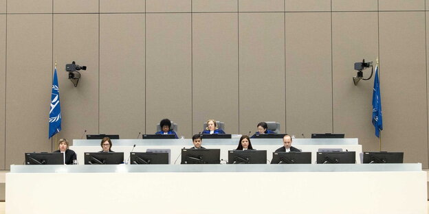 Ein Gerichtssaal mit beigen Wänden und einem langen Tisch, an dem Menschen sitzen Ein Gerichtssaal mit beigen Wänden und einem langen Tisch, an dem Menschen sitzen