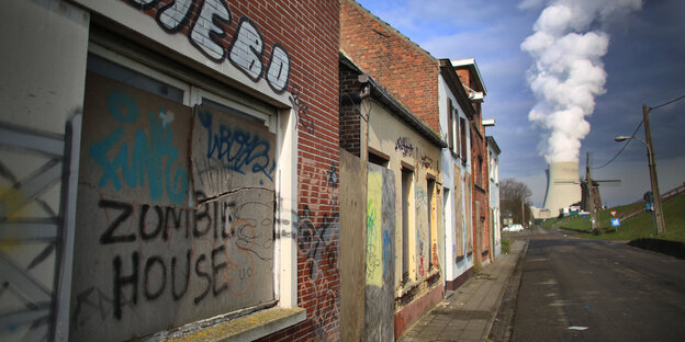 Eine verlassene Häuserreihe, auf einem Brett steht „Zombie House“. Am Ende der Straße ein Kühlturm, aus dem Dampf aufsteigt Eine verlassene Häuserreihe, auf einem Brett steht „Zombie House“. Am Ende der Straße ein Kühlturm, aus dem Dampf aufsteigt