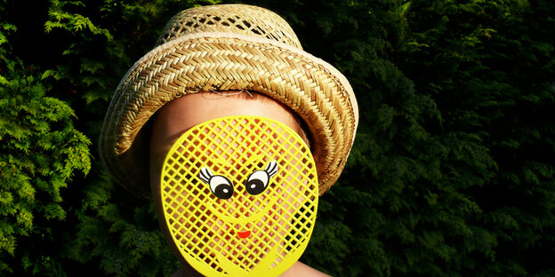 Eine Person mit Hut hält sich eine gelbe Fliegenklatsche vor ihr Gesicht, auf der Klatsche ist ein Smiley Eine Person mit Hut hält sich eine gelbe Fliegenklatsche vor ihr Gesicht, auf der Klatsche ist ein Smiley