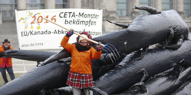 Frau sticht mit Mistgabel in schwarze Gummiwülste, die ein Monster darstellen, dahinter ein Transparent mit der Aufschrift "Ceta-Monster bekämpfen" Frau sticht mit Mistgabel in schwarze Gummiwülste, die ein Monster darstellen, dahinter ein Transparent mit der Aufschrift "Ceta-Monster bekämpfen"