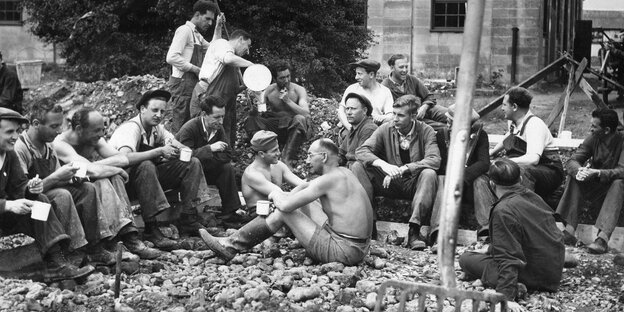 Ein Schwarz-Weiß-Foto, darauf Männer, die inmitten von Schutt sitzen und Pause machen Ein Schwarz-Weiß-Foto, darauf Männer, die inmitten von Schutt sitzen und Pause machen