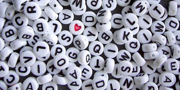Weiße Perlen mit schwarzen Buchstaben auf einem Haufen, dazwischen eine Perle mit rotem Herz Weiße Perlen mit schwarzen Buchstaben auf einem Haufen, dazwischen eine Perle mit rotem Herz