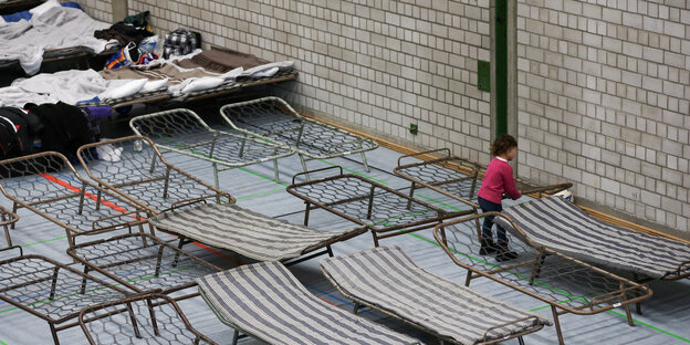 Feldbetten stehen in einer Turnhalle, dazwischen ein Mädchen Feldbetten stehen in einer Turnhalle, dazwischen ein Mädchen