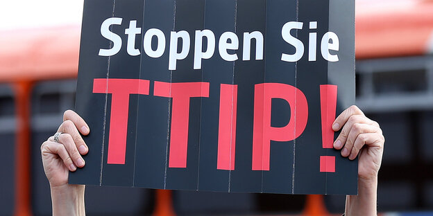 Zwei Hände halten ein schwarzes Schild, auf dem in weißer Schrift "Stoppen Sie" und in schwarzer "TTIP" steht Zwei Hände halten ein schwarzes Schild, auf dem in weißer Schrift "Stoppen Sie" und in schwarzer "TTIP" steht