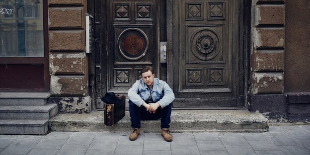 Ein Mann sitzt vor einer Haustür, neben sich einen Koffer, darauf ein Hut Ein Mann sitzt vor einer Haustür, neben sich einen Koffer, darauf ein Hut