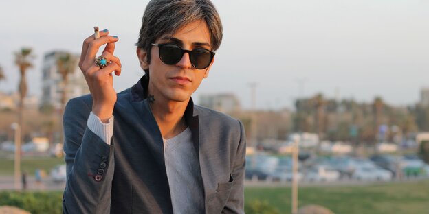 Payam Feili sitzt mit Sonnenbrille, Zigarette in der Hand, blau lakierten Fingernägeln und einem blauen Ring vor einer Stadtlandschaft. Am Hals ein Davidstern-Tattoo Payam Feili sitzt mit Sonnenbrille, Zigarette in der Hand, blau lakierten Fingernägeln und einem blauen Ring vor einer Stadtlandschaft. Am Hals ein Davidstern-Tattoo