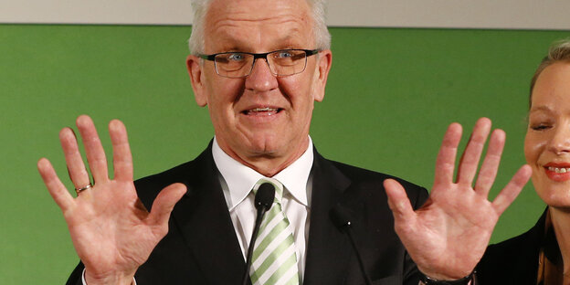 Ein Mann, es ist Winfried Kretschmann, hebt die Hände in die Luft und steht vor einem grünen Hintergrund Ein Mann, es ist Winfried Kretschmann, hebt die Hände in die Luft und steht vor einem grünen Hintergrund