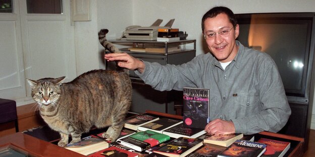 Akif Pirinçci am Schreibtisch sitzend. Er präsentiert eines seiner Bücher und streichelt eine Katze, die sich auf dem Tisch befindet. Akif Pirinçci am Schreibtisch sitzend. Er präsentiert eines seiner Bücher und streichelt eine Katze, die sich auf dem Tisch befindet.