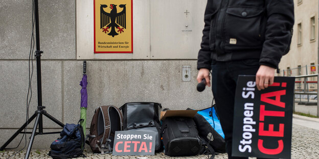Einige Protestutensilien, ein Lautsprecherständer und ein mensch mit einem Anti-Ceta-Schild stehen vor einer grauen Mauer Einige Protestutensilien, ein Lautsprecherständer und ein mensch mit einem Anti-Ceta-Schild stehen vor einer grauen Mauer