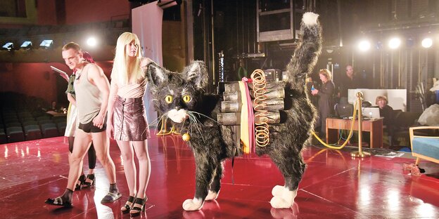 Eine Frau im Minirock steht mit einem Mann auf der Theaterbühne, neben ihr eine überlebensgroße Katze mit Sprengstoffgürtel Eine Frau im Minirock steht mit einem Mann auf der Theaterbühne, neben ihr eine überlebensgroße Katze mit Sprengstoffgürtel