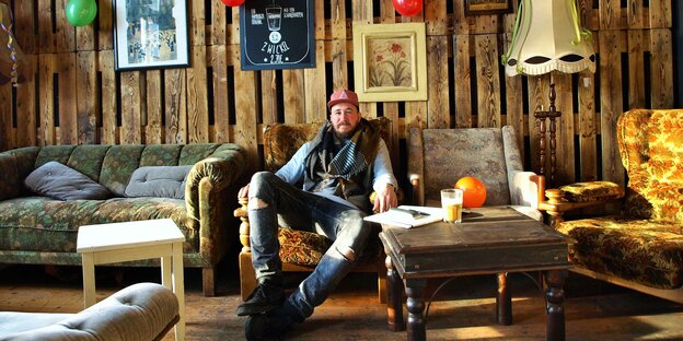 Keno Hellmers sitzt auf einen Sofa vor einer Wand aus Europaletten. Keno Hellmers sitzt auf einen Sofa vor einer Wand aus Europaletten.