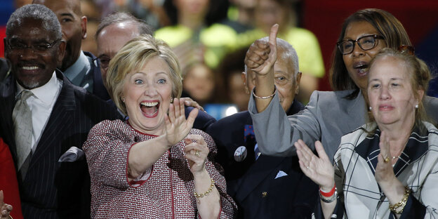 Hillary Clinton winkt und jubelt, umringt von klatschenden UnterstützerInnen Hillary Clinton winkt und jubelt, umringt von klatschenden UnterstützerInnen