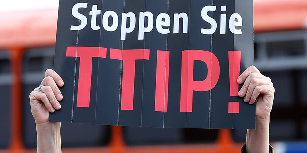 Zwei Hände halten ein schwarzes Schild mit der Aufschrift „Stoppen Sie TTIP!“ in die Luft Zwei Hände halten ein schwarzes Schild mit der Aufschrift „Stoppen Sie TTIP!“ in die Luft
