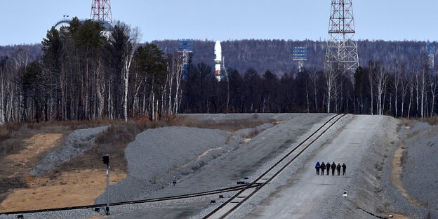 einige Menschen laufen neben Gleisen, im Hintergrund eine Rakete und Wald einige Menschen laufen neben Gleisen, im Hintergrund eine Rakete und Wald
