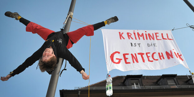 Eine Frau hängt kopfüber an einem Fahnenmast neben einem Banner mit der Aufschrift "Kriminell ist die Gentechnik" Eine Frau hängt kopfüber an einem Fahnenmast neben einem Banner mit der Aufschrift "Kriminell ist die Gentechnik"