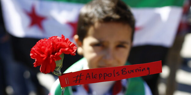 Ein Kind mit Nelke und dem Spruchband „Aleppo is burning“ vor einer syrischen Flagge Ein Kind mit Nelke und dem Spruchband „Aleppo is burning“ vor einer syrischen Flagge