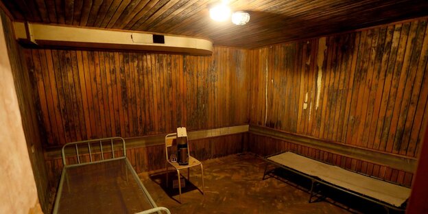 Ein holzvertäfelter Raum, darin ein grünliches Bettgestell aus Eisen, ein Stuhl und eine Pritsche Ein holzvertäfelter Raum, darin ein grünliches Bettgestell aus Eisen, ein Stuhl und eine Pritsche