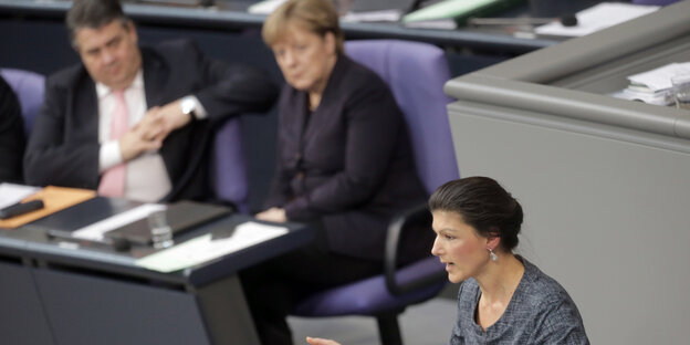 Sarah Wagenknecht spricht im Bundestag. Sigmar Gabriel und Angela Merkel sitzen im Hintergrund und hören zu. Sarah Wagenknecht spricht im Bundestag. Sigmar Gabriel und Angela Merkel sitzen im Hintergrund und hören zu.