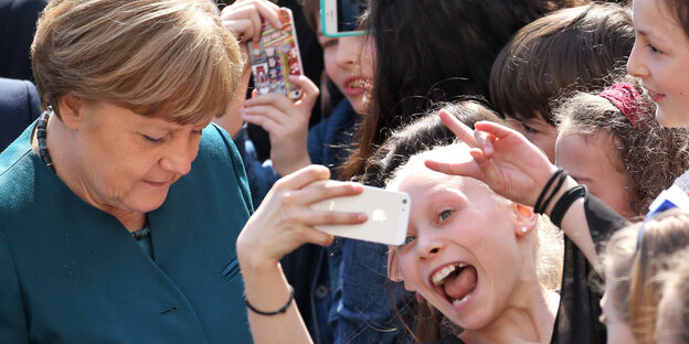 Merkel guckt nach unten, vor ihr eine Schülerin, die irr in ein zum Selfie gezücktes Handy grinst Merkel guckt nach unten, vor ihr eine Schülerin, die irr in ein zum Selfie gezücktes Handy grinst
